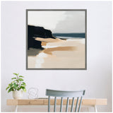 Abstract coastal landscape painting with soft sand tones, dark cliff contrasts, and muted gray sky, framed and hung above a light wood desk with a plant and chair.