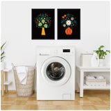 Two framed floral artworks with black backgrounds above a white front-load washing machine, featuring stylized yellow and red vases with colorful flowers.