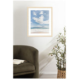 Framed beach photo showing white sand, turquoise ocean, and a cloudy blue sky, hanging on a white wall above a dark green armchair and potted plant.