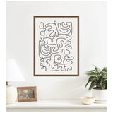 Framed abstract canvas print with continuous black line shapes on white background, displayed on a white wall above a table with a lamp, photo frame, and potted plant.