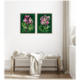 Two framed floral paintings with pink and light green abstract flowers on dark green backgrounds, hung side by side above a wooden bench with a draped white cloth and open book.