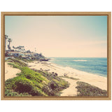 Sandy coastal beach with green vegetation leading to calm blue ocean waves, bordered by houses and palm trees under a clear sky in soft vintage tones.