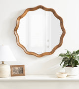 Round mirror with a scalloped wooden frame featuring smooth curves, hanging above a white shelf with neutral-toned lamp, photo frame, and potted green plant.