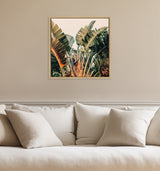 Framed canvas print of large green tropical banana leaves with reddish-brown stems hanging above a beige sofa with neutral cushions.