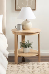 Round light wood side table with a lower shelf, holding a beige ceramic table lamp and a gray mug on top, plus a green plant in a cream pot below.