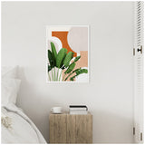 Framed abstract botanical canvas with large green leaves against terracotta and beige geometric shapes, hanging on a white wall above a rustic wooden side table.