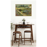 Vincent van Gogh's Garden at Arles print with vibrant green, blue, and yellow garden scene, hung above a wooden bar table with stools and minimalist tableware.