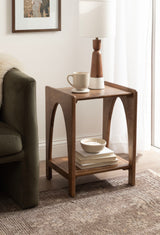 Mid-century modern wooden side table with curved arches on legs, topped with a white and wood table lamp and a beige mug, beside a green sofa.