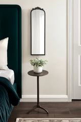 Tall, narrow arch mirror with a black ornate frame hangs above a small round black metal side table holding a white pot with green eucalyptus branches, set against a beige wall.