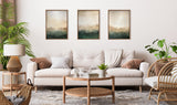 Living room with a white sofa adorned with brown and patterned pillows, a wicker coffee table, three framed green and beige abstract mountain art pieces, and plants on either side.