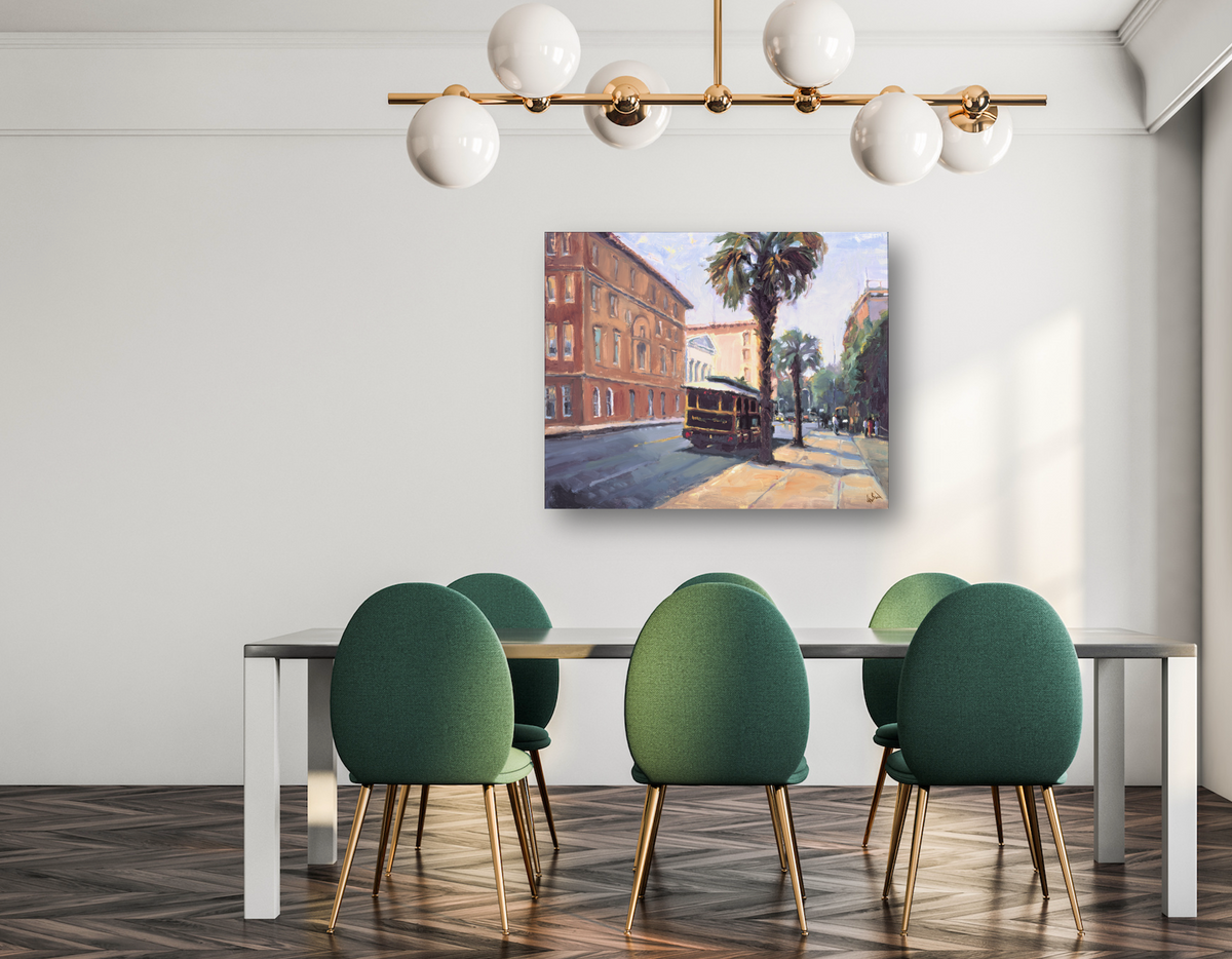 Canvas print of a Charleston street scene featuring a trolley, palm trees, and historic brick buildings, hung above a modern dining table with green chairs.