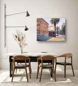 Wall-mounted canvas painting showing a vintage trolley on a sunlit urban street with palm trees and classic brick buildings in warm tones.