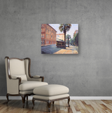 Canvas print of a street scene with a vintage trolley, palm trees, and sunlit sidewalks, displayed on a gray wall above a cream armchair with wooden legs and matching ottoman.