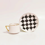 White espresso cup with gold rim and handle alongside a matching saucer featuring a black and white checkered pattern with gold rim.
