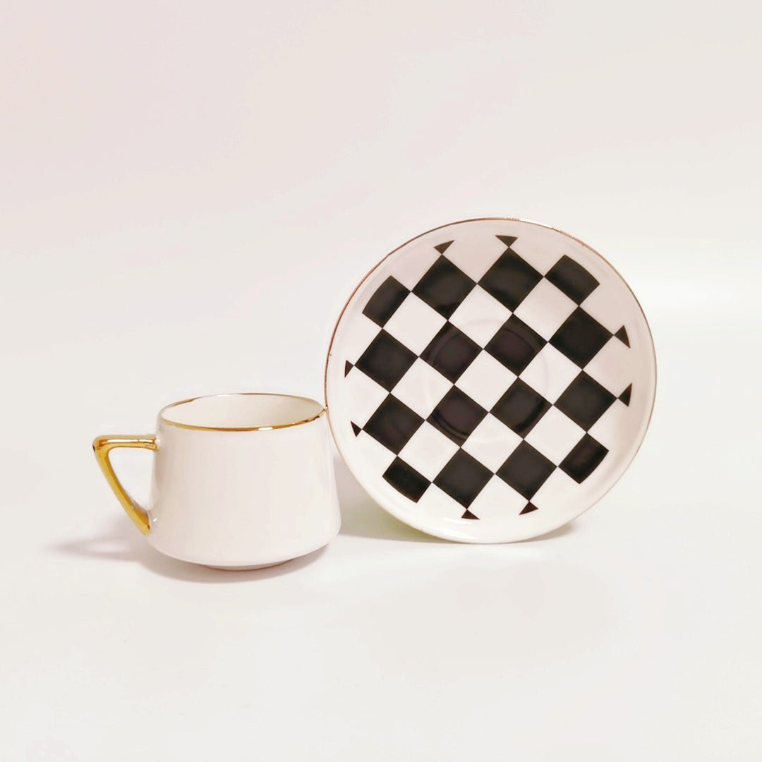 White espresso cup with gold rim and handle alongside a matching saucer featuring a black and white checkered pattern with gold rim.