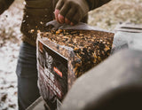 Close-up of a hand wearing a brown glove holding a knife cutting into a rectangular block of fortified deer bait with seeds and grains embedded on top, labeled BIG TINE FORTIFIED DEER BLOCK CHERRY.