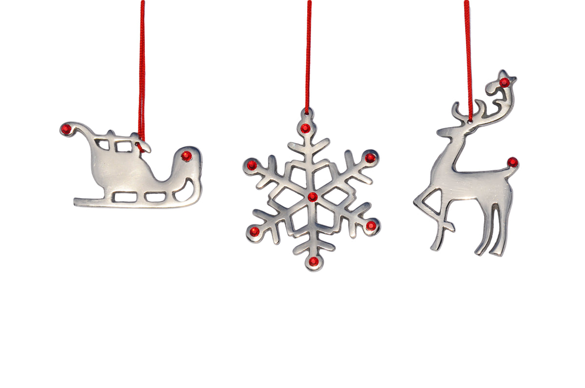 Three silver Christmas ornaments shaped as a sleigh, snowflake, and reindeer, each adorned with small red ruby dots and hanging from red strings.