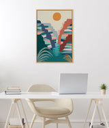 Abstract canyon landscape in muted blues, greens, reds, and pinks with an orange sun above a teal river, framed and hanging on a white wall over a modern desk.