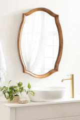 Wall mirror with a scalloped natural wood frame above a white countertop featuring a round vessel sink, gold faucet, and small green potted plant.