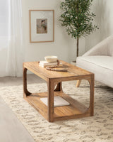 Rectangular wooden coffee table with curved edges and open lower shelf, displayed on a patterned beige rug beside a light gray sofa and green potted plant.