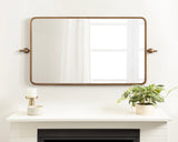Rectangular metal-framed mirror with rounded corners and bronze finish, mounted above a white mantel holding a plant in a beige pot and a white candle holder.
