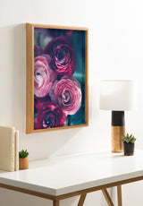 Framed art print of pink and deep purple ranunculus flowers with green-blue background, hanging on a white wall above a white desk with a lamp and small succulent plants.