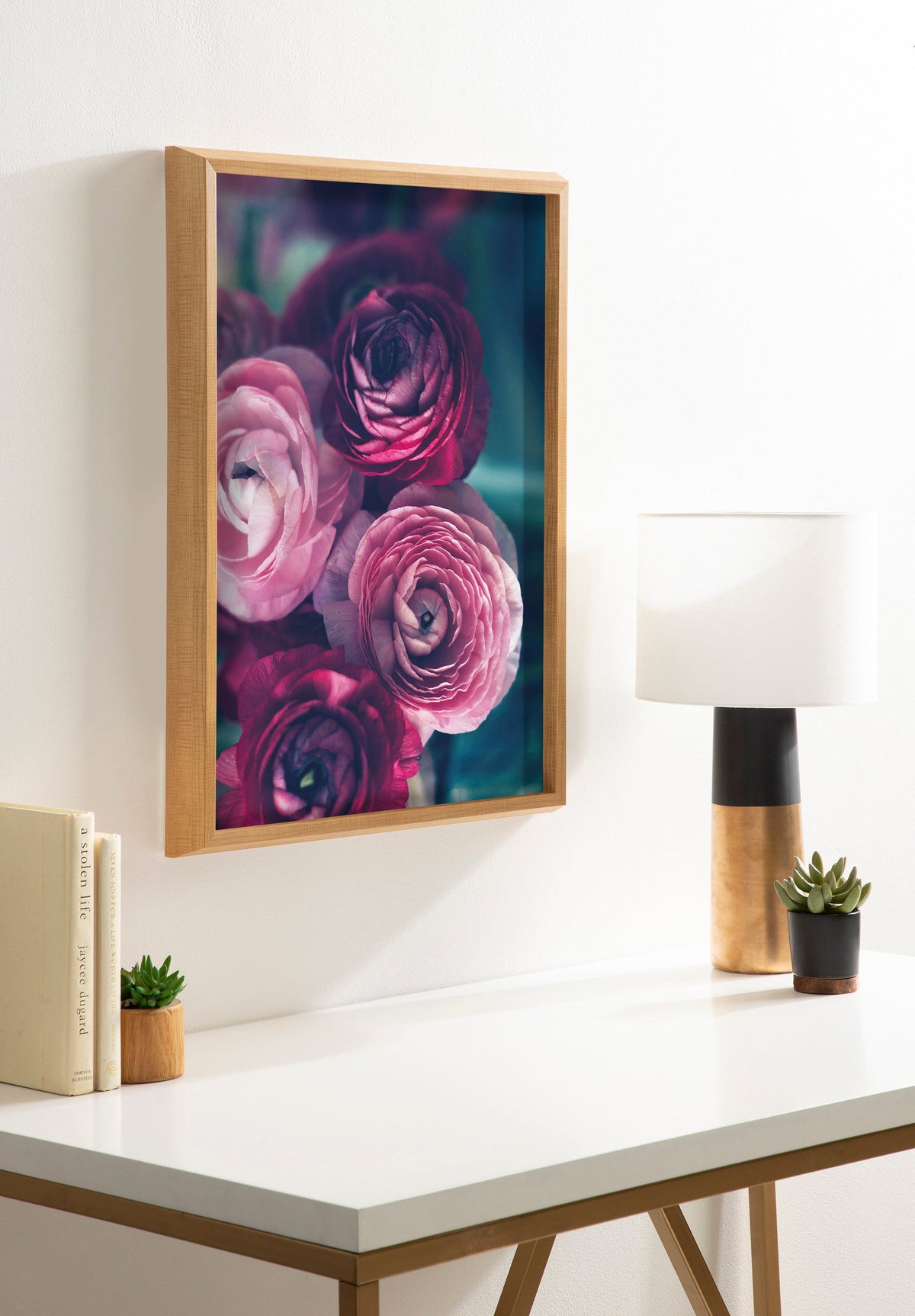 Framed art print of pink and deep purple ranunculus flowers with green-blue background, hanging on a white wall above a white desk with a lamp and small succulent plants.