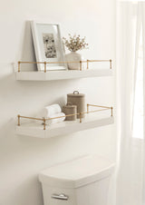 Two white floating shelves with gold metal railings mounted on a cream wall, decorated with neutral-toned jars, rolled white towels, a flower vase, and a framed black-and-white botanical print.