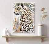 Framed canvas artwork of a cheetah with detailed spots and blue eyes, surrounded by beige and green tropical leaves, mounted on a wall above a wooden shelf.