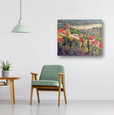 Impressionist-style painting of a clock tower amidst red-roofed buildings and green trees, hanging on a white wall above a wooden armchair with green cushions.