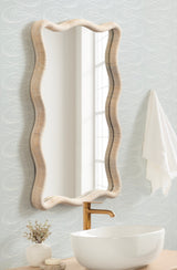 Rectangular wall mirror with a scalloped, wavy rattan frame hanging above a white vessel sink and gold faucet against light gray wallpaper with subtle leaf patterns.