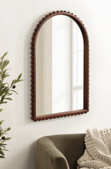 Arched wall mirror with a dark brown frame featuring rounded bead-like trim, hung on a white wall above a green sofa with a textured cream throw blanket.