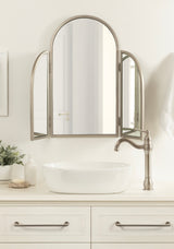 Tri-fold metal mirror with arched top mounted above white oval vessel sink and beige countertop, flanked by brushed nickel faucet and soap dispenser.