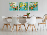 Three framed canvas paintings of fruit on branches with green leaves against blue backgrounds, hung in a row above a modern dining table with white chairs and wooden accents.