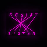 Rectangular black sign with neon pink text 'RESIST' and 'SISTER' connected by multiple diagonal neon lines forming a starburst pattern.