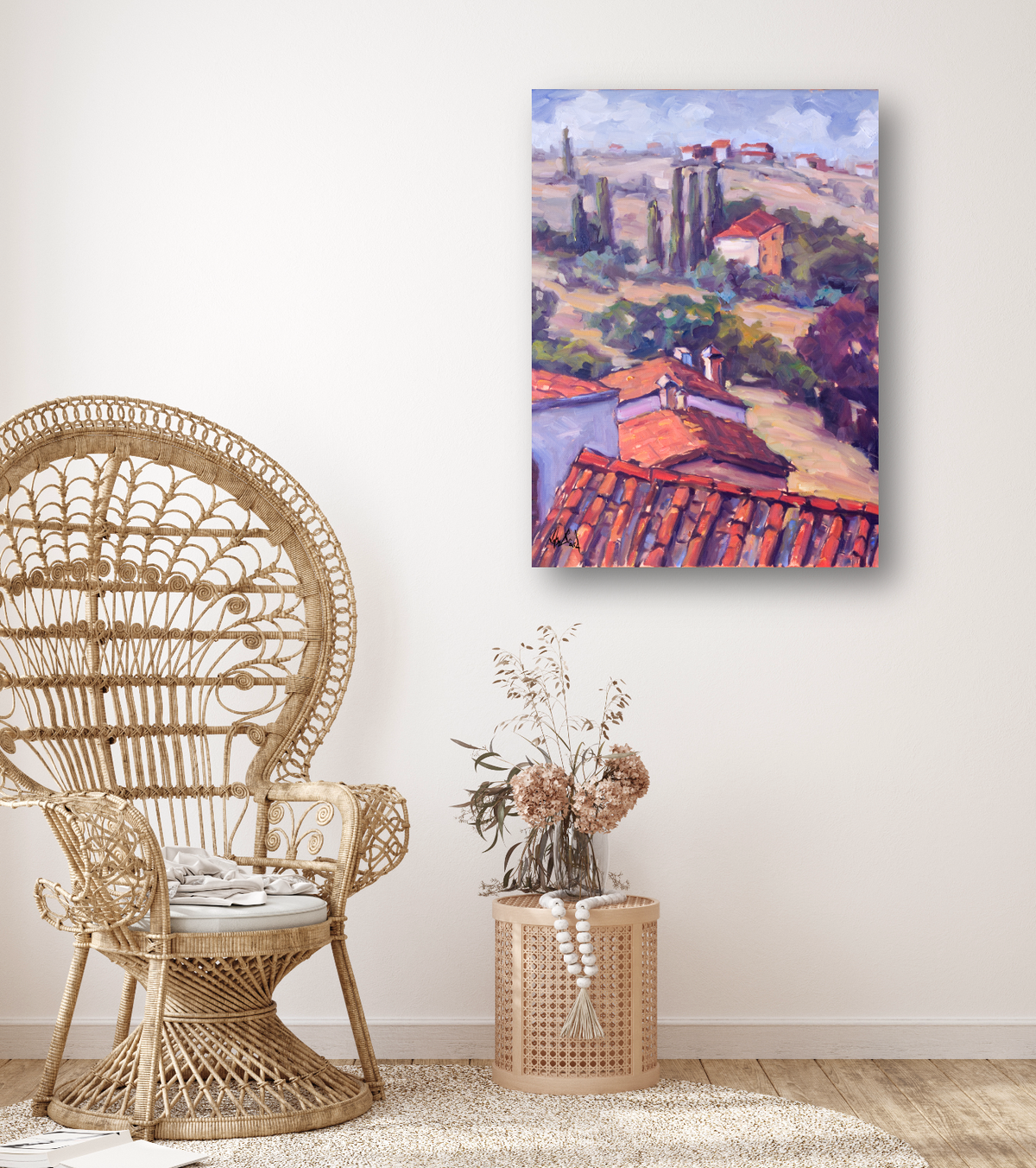 Canvas print of a landscape with red-tiled rooftops and cypress trees in soft, impressionistic brushstrokes, hung on a white wall above a rattan chair and side table with dried flowers.