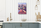 Small colorful canvas print of Cortona landscape featuring terracotta rooftops and distant cypress trees, hung on white paneled wall above pale blue kitchen cabinets.