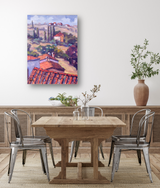 Impressionist-style canvas painting of Cortona landscape with terracotta roofs, cypress trees, and rolling hills in warm purples and reds, hung above a wooden dining table.