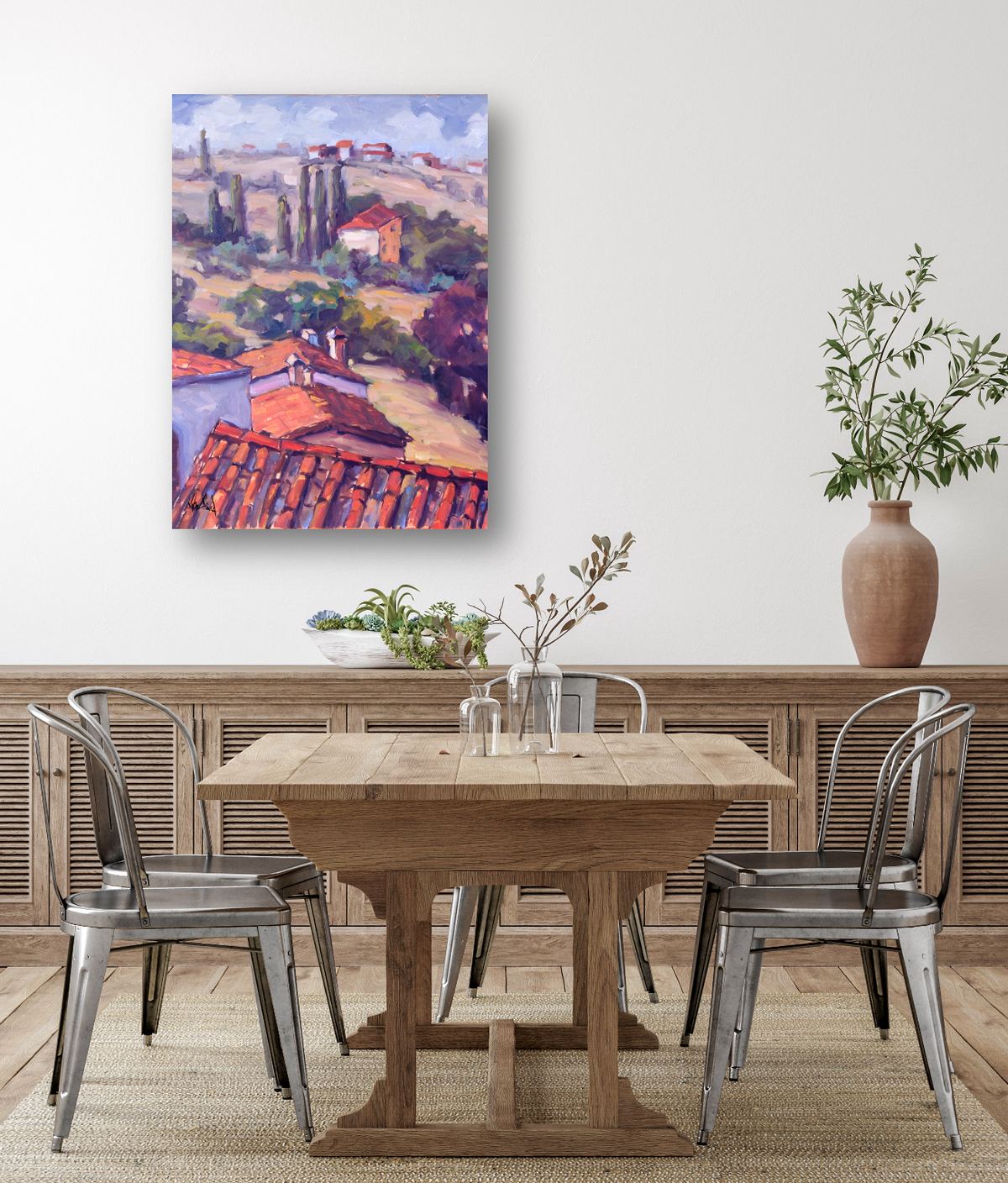 Impressionist-style canvas painting of Cortona landscape with terracotta roofs, cypress trees, and rolling hills in warm purples and reds, hung above a wooden dining table.