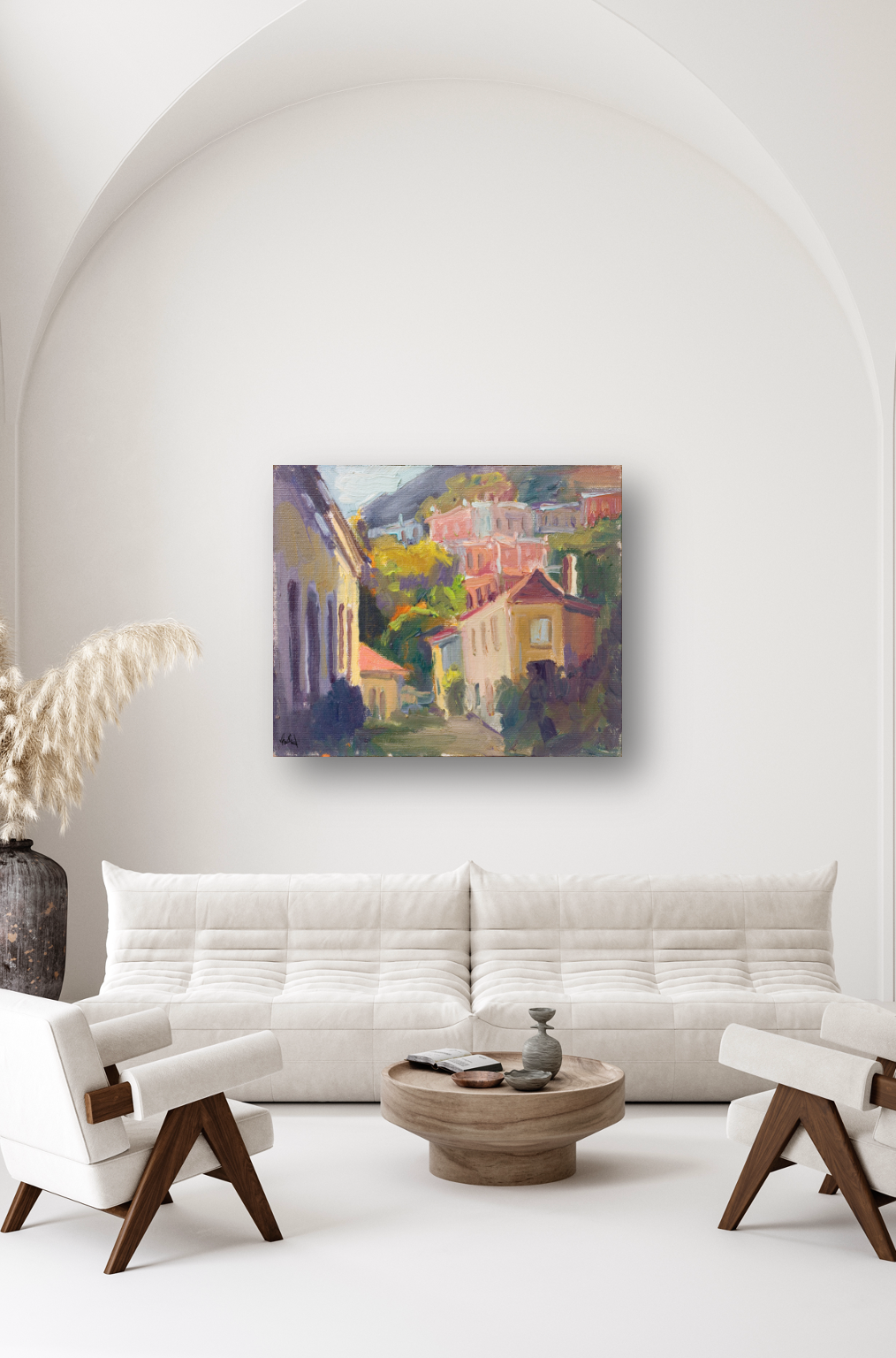 Impressionist-style canvas print of colorful hillside cottages with trees and sky, mounted on a white arched wall above a modern cream sofa and wood chairs.