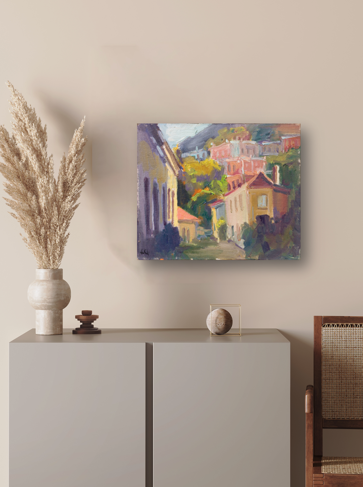 Impressionist-style painting of hillside cottages with warm yellow and pink tones, hung above a modern beige cabinet decorated with pampas grass and minimalist objects.