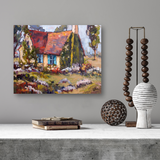 Impressionist canvas painting of a small cottage with a red roof and bright blue shutters surrounded by greenery and flowers, hung on a gray wall above a concrete surface with decorative objects.