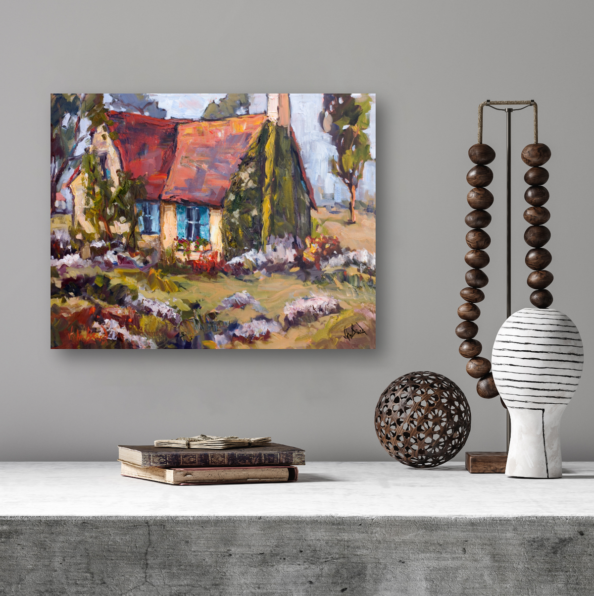 Impressionist canvas painting of a small cottage with a red roof and bright blue shutters surrounded by greenery and flowers, hung on a gray wall above a concrete surface with decorative objects.