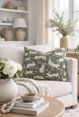 Square sage green pillow with white cottontail rabbit silhouettes on a light beige sofa, next to a cream throw and neutral-toned decor including a textured vase with greenery.