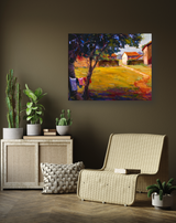 Colorful painting of a sunlit country path leading past cottages with a prominent tree casting shadows in the foreground, displayed on a brown wall above a wicker chair and plants.