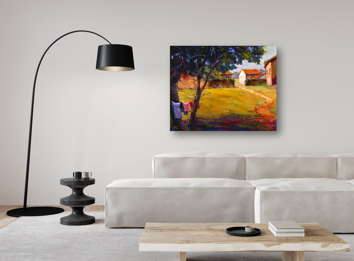 Bright canvas print of a country cottage scene with a yellow path and lush green and purple trees, displayed above a modern light beige sofa in a minimalist room.
