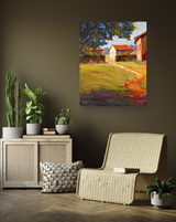 Canvas print of a sunlit country scene with a dirt path leading to white and red-roofed houses, displayed above a rattan chair and cabinet with green plants.