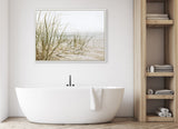 Minimalist bathroom with a white oval freestanding tub beneath a framed canvas depicting sandy beach grass in muted natural tones, next to wooden shelving with towels.