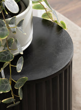 Close-up of a round black side table with vertical ridges along the sides, topped with a white plant pot and trailing green leaves.