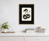 Black and cream graphic of two cherries with billiard 8-ball patterns hanging from a stem with leaves, framed in natural wood and hung on a white wall above a white cabinet.